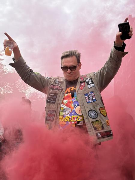 A man with sunglasses and a patched jacket raises his arms in celebration amidst a cloud of vibrant red smoke